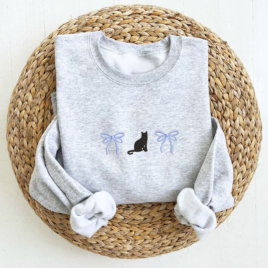 Gray sweatshirt with a black cat and periwinkle bow embroidered design folded on top of a woven mat.