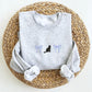 Gray sweatshirt with a black cat and periwinkle bow embroidered design folded on top of a woven mat.