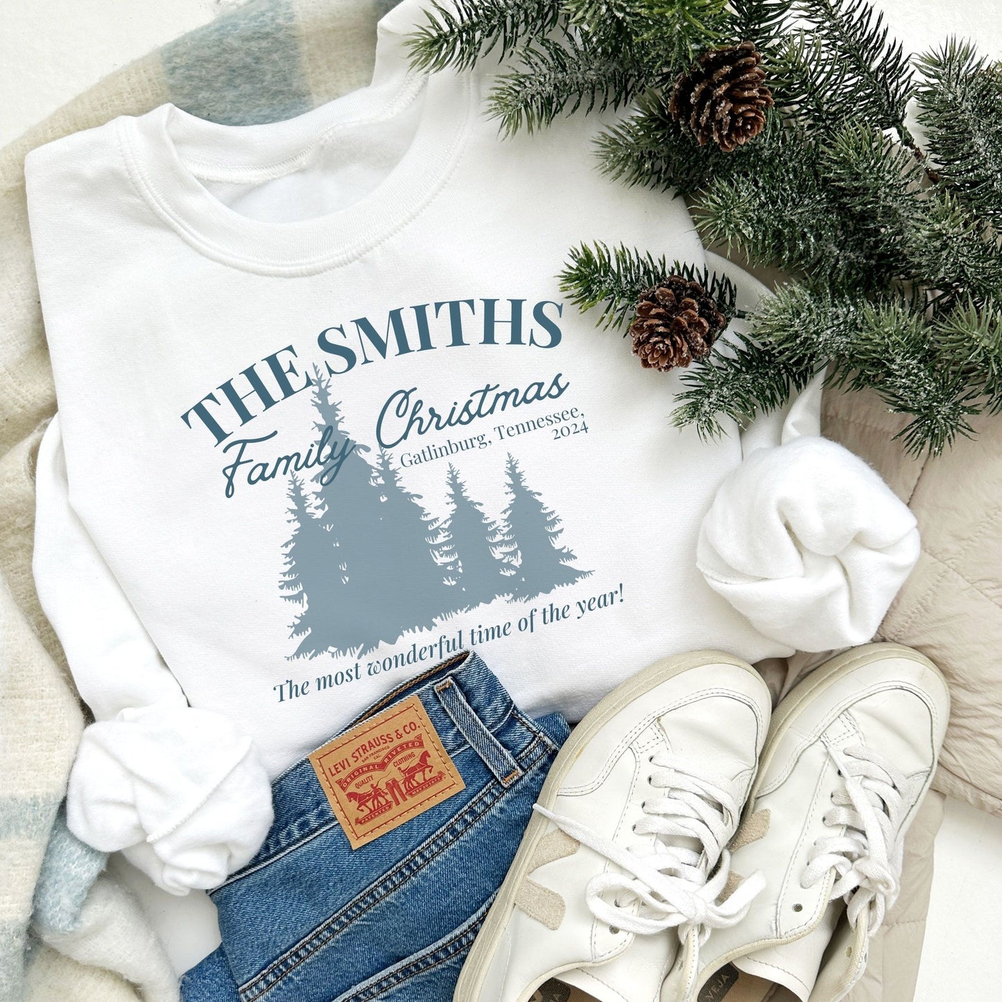 A white crewneck sweatshirt with a custom Christmas print featuring a family name, vacation location, year, trees, and a "The most wonderful time of the year!" quote in blue ink