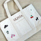 White tote bag with embroidered designs and text on a wooden surface