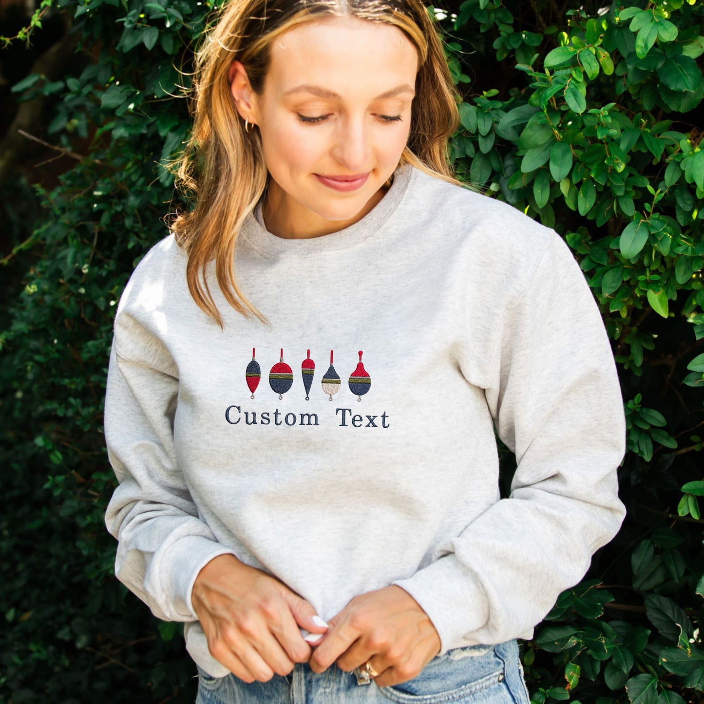 A woman wearing a light gray crewneck sweatshirt with a graphic of four fishing bouys and the text 'Custom Text' printed on the chest, set against a background of green foliage.