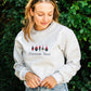A woman wearing a light gray crewneck sweatshirt with a graphic of four fishing bouys and the text 'Custom Text' printed on the chest, set against a background of green foliage.