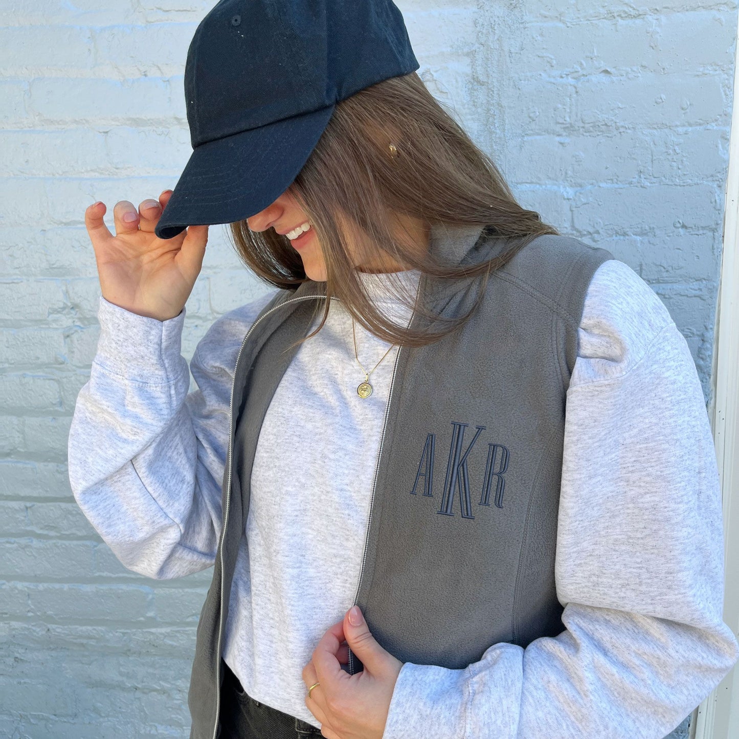 young woman wearing a lightweight grey fleece vest with a navy blue embroidered monogram on the left chest