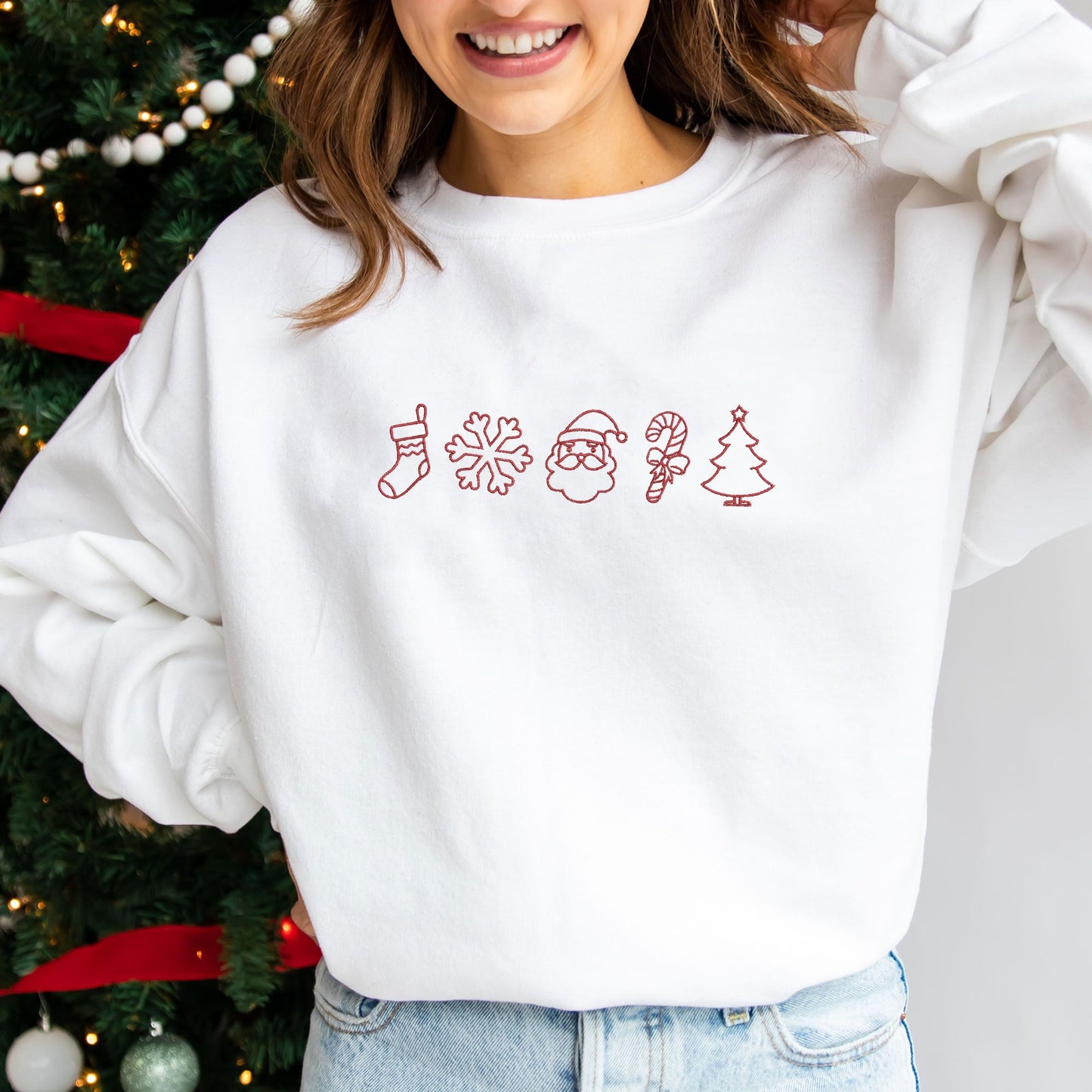 young woman modeling a white crewneck sweatshirt with christmas outlined icons embroidered across the chest in red thread.