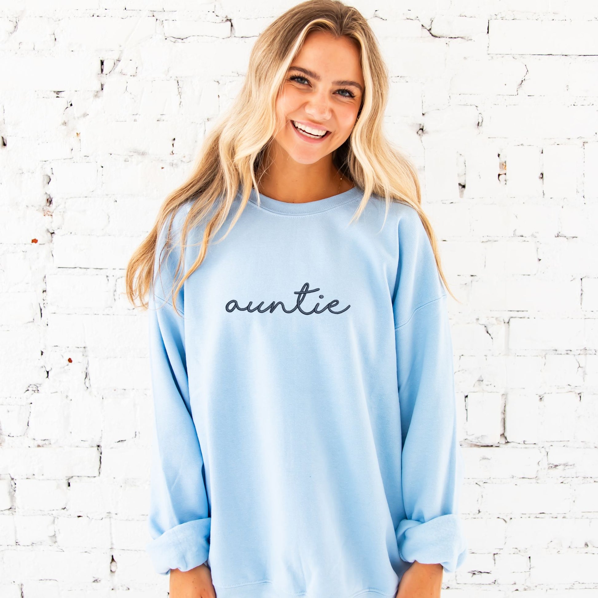 young woman wearing an oversize light blue crewneck sweatshirt with embroidered auntie in lowercase script font across the chest in navy thread