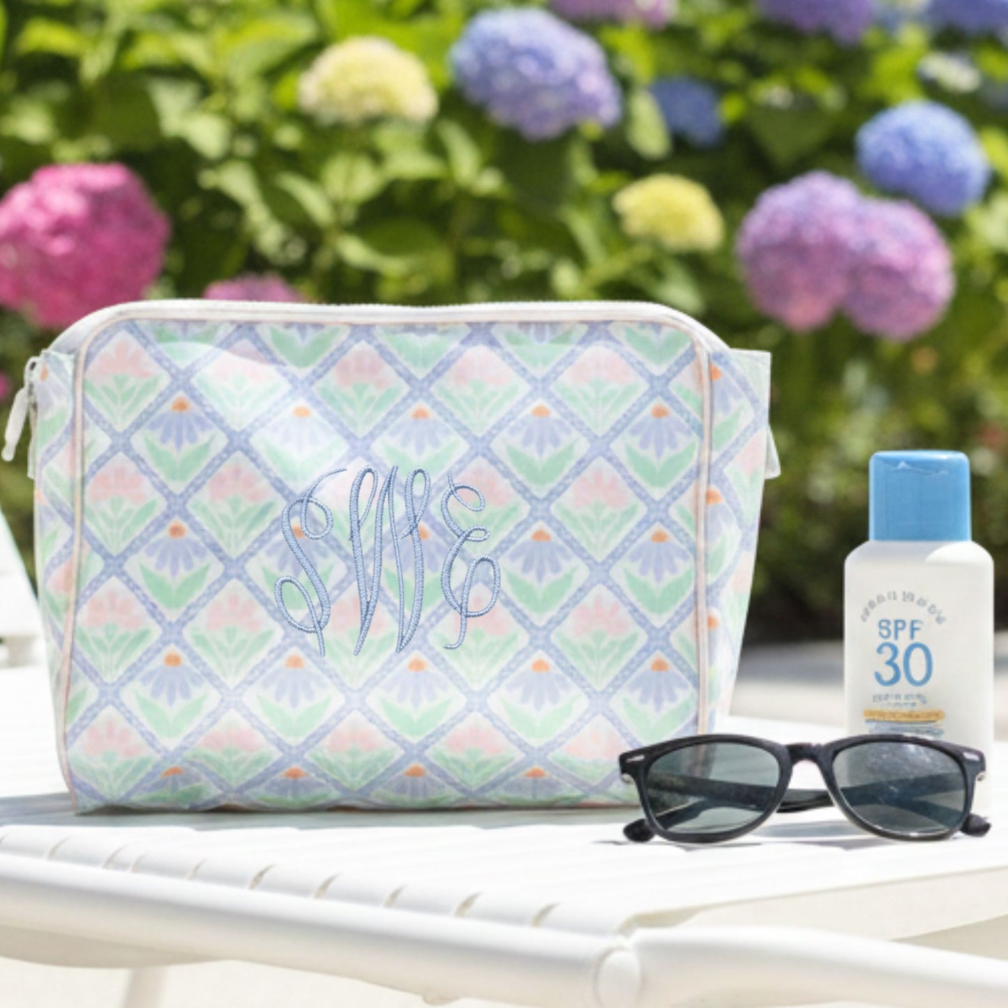 Patterned toiletry bag with sunglasses and sunscreen on a table with flowers in the background