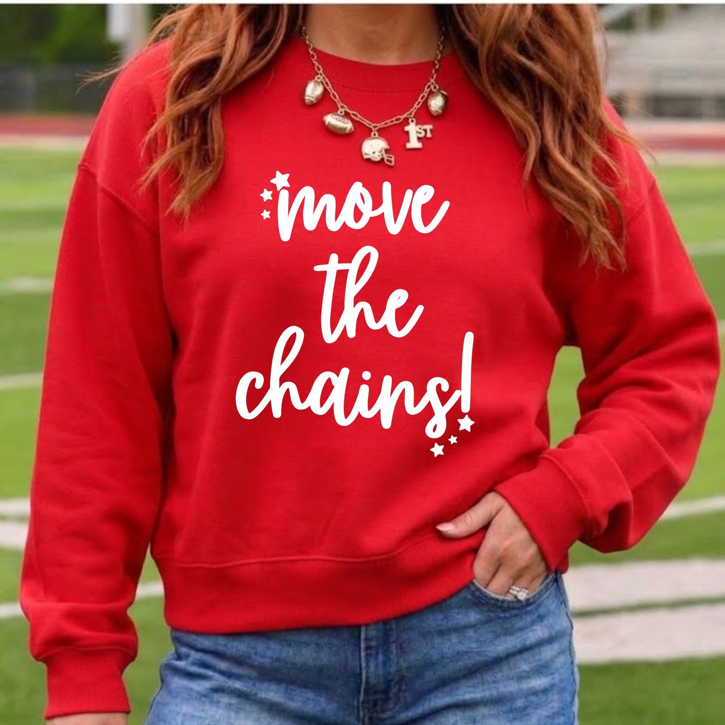 A woman wearing a bright red crewneck sweatshirt with the phrase 'move the chains!' printed in white script across the front, paired with blue jeans and a gold necklace, set against a blurred background of a sports field.
