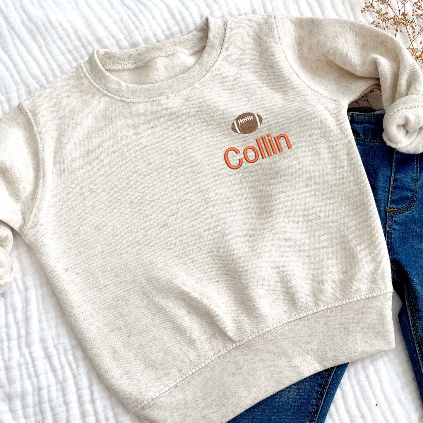 a natural heather crewneck toddler sweatshirt with custom name embroidery on the left chest featuring a mini football above the name