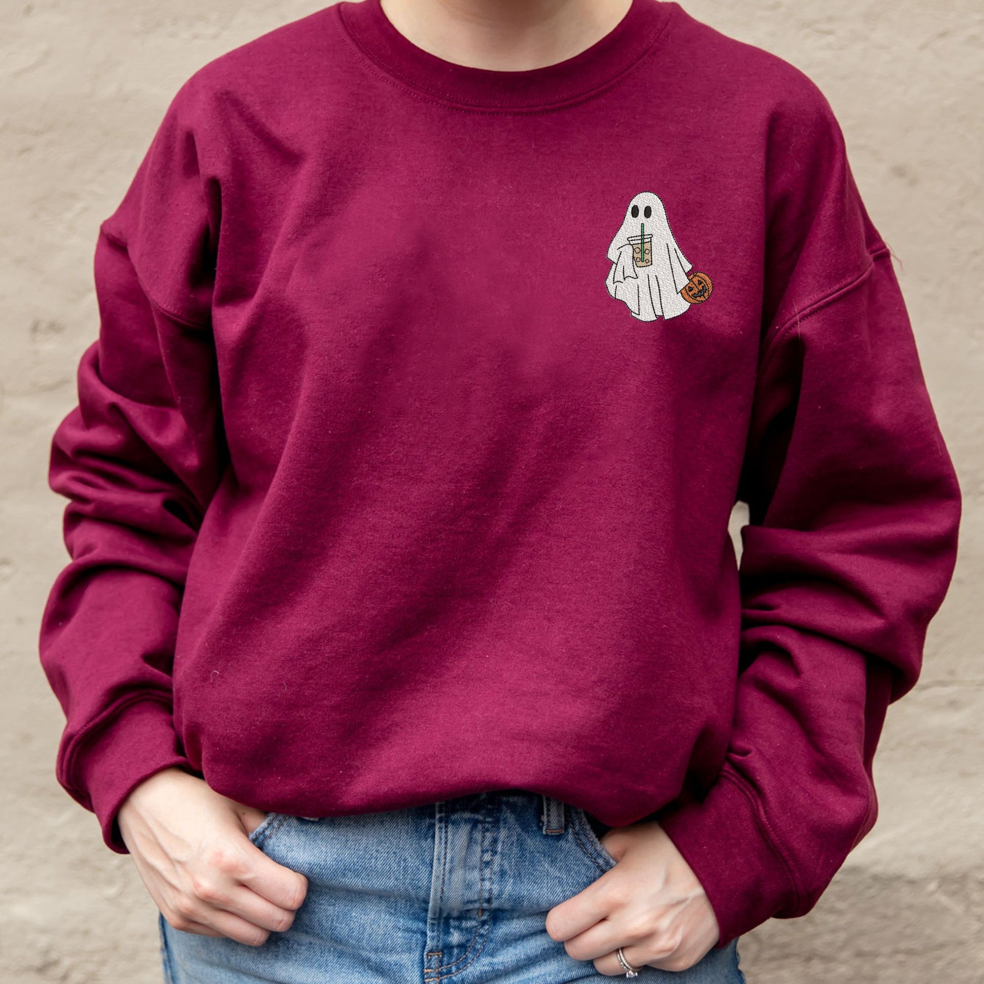 close up of a maroon crewneck sweatshirt with embroidered ghost drinking iced coffee and holding a jack-o-lantern on the left chest