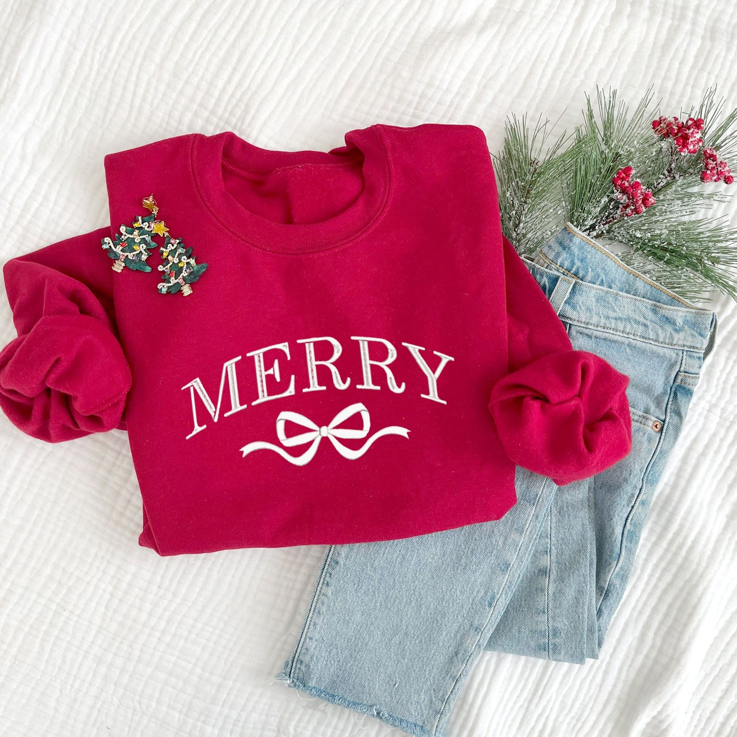 Christmas styled flat lay of a cardinal red crewneck with embroidered merry and bow design in white  thread