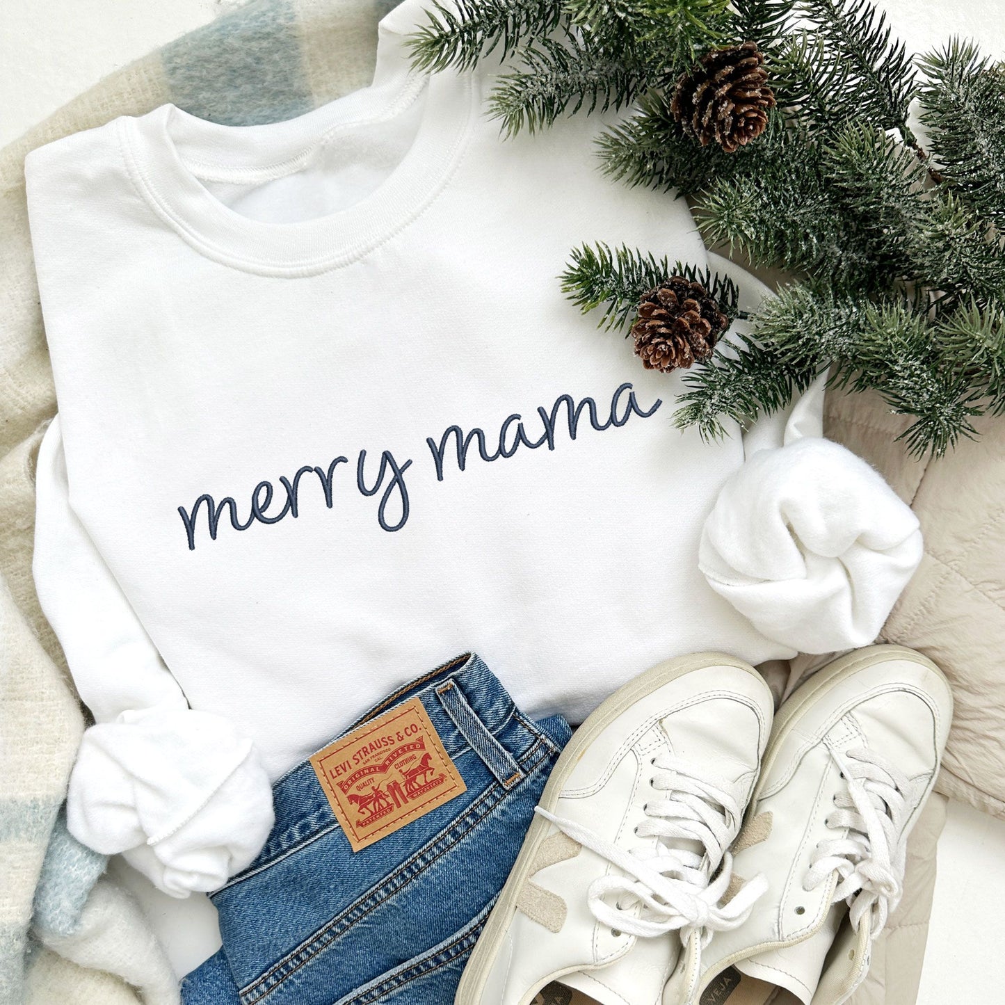 white crewneck sweatshirt with embroidered merry mama in navy thread styled with Christmas decor, jeans, and sneakers