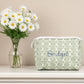Green patterned pouch with 'Bridget' embroidery next to a vase of white daisies on a beige background