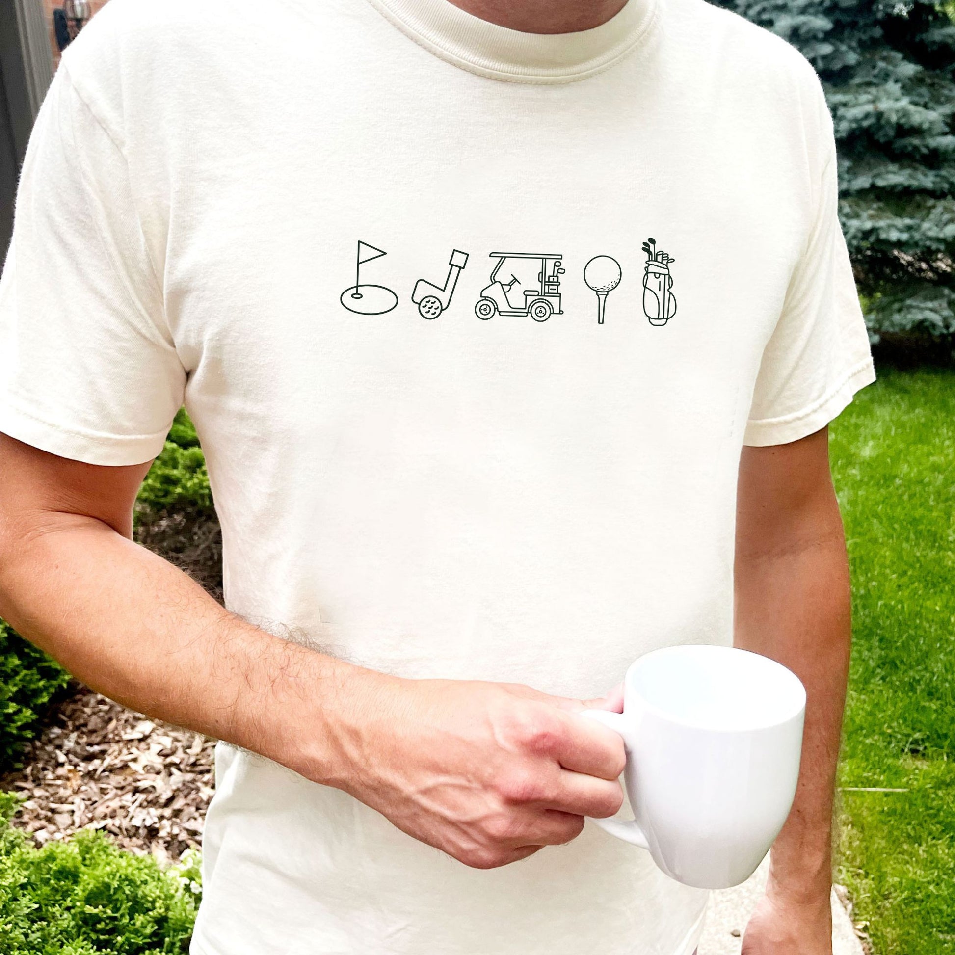 man standing outside wearing an ivory comfort colors t-shirt with custom outlined golf icon printed design featuring: golf bag, golf ball on a tee, golf cart, golf club, and golf green/flag