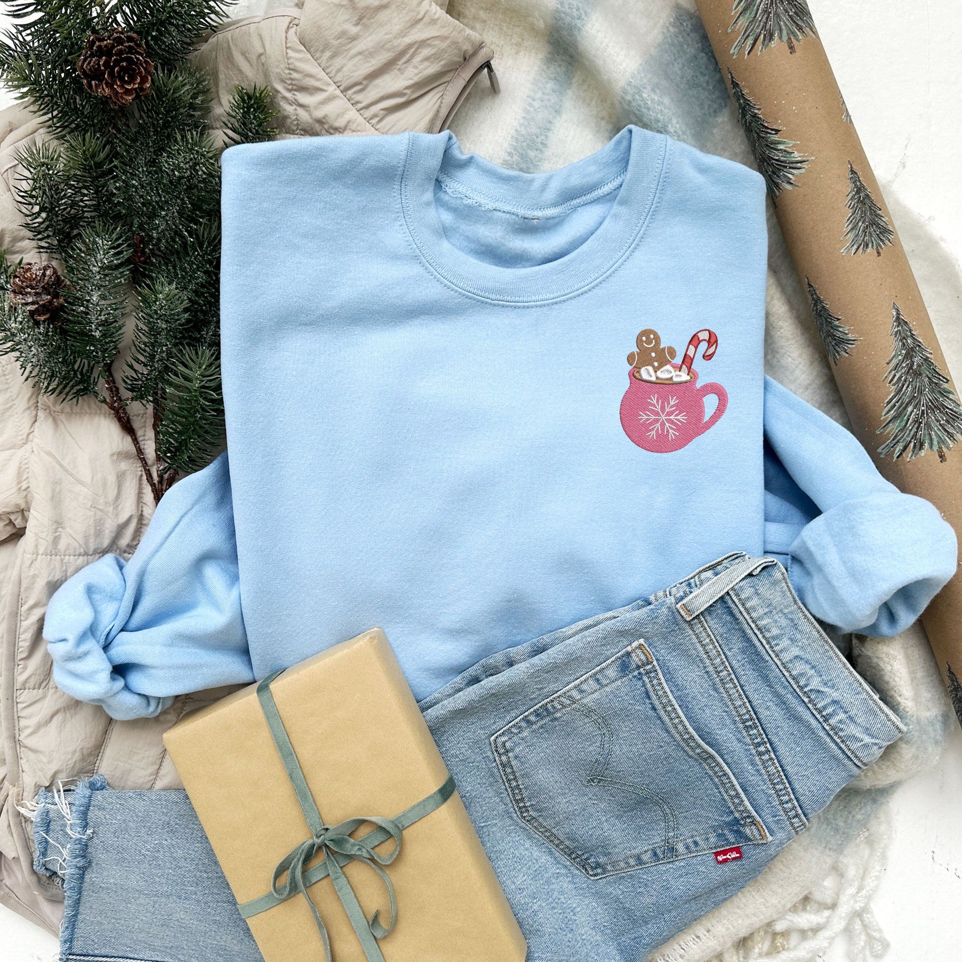 festive Christmas flat lay photo of a light blue crewneck sweatshirt with embroidered pink mug with marshmallows a a ginger breadman and candy cane on the left chest.