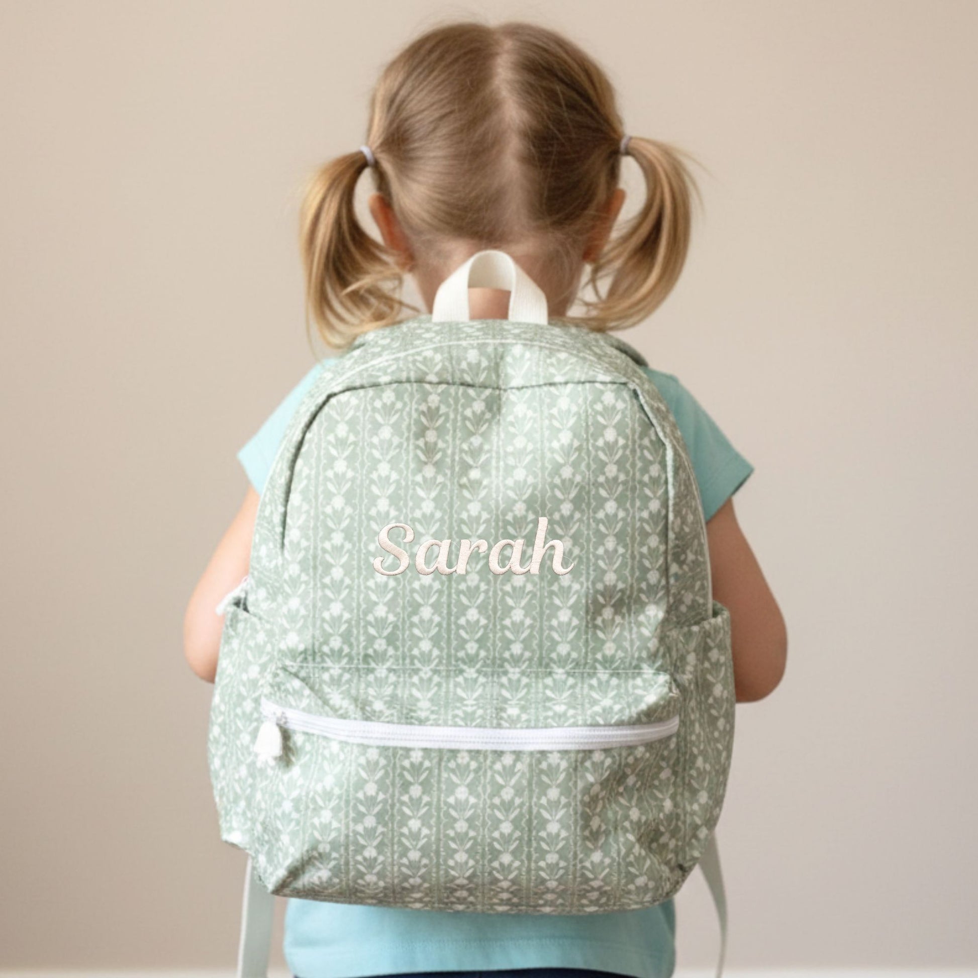 Child wearing a sage green floral patterned backpack with 'Sarah' embroidered on it against a beige background