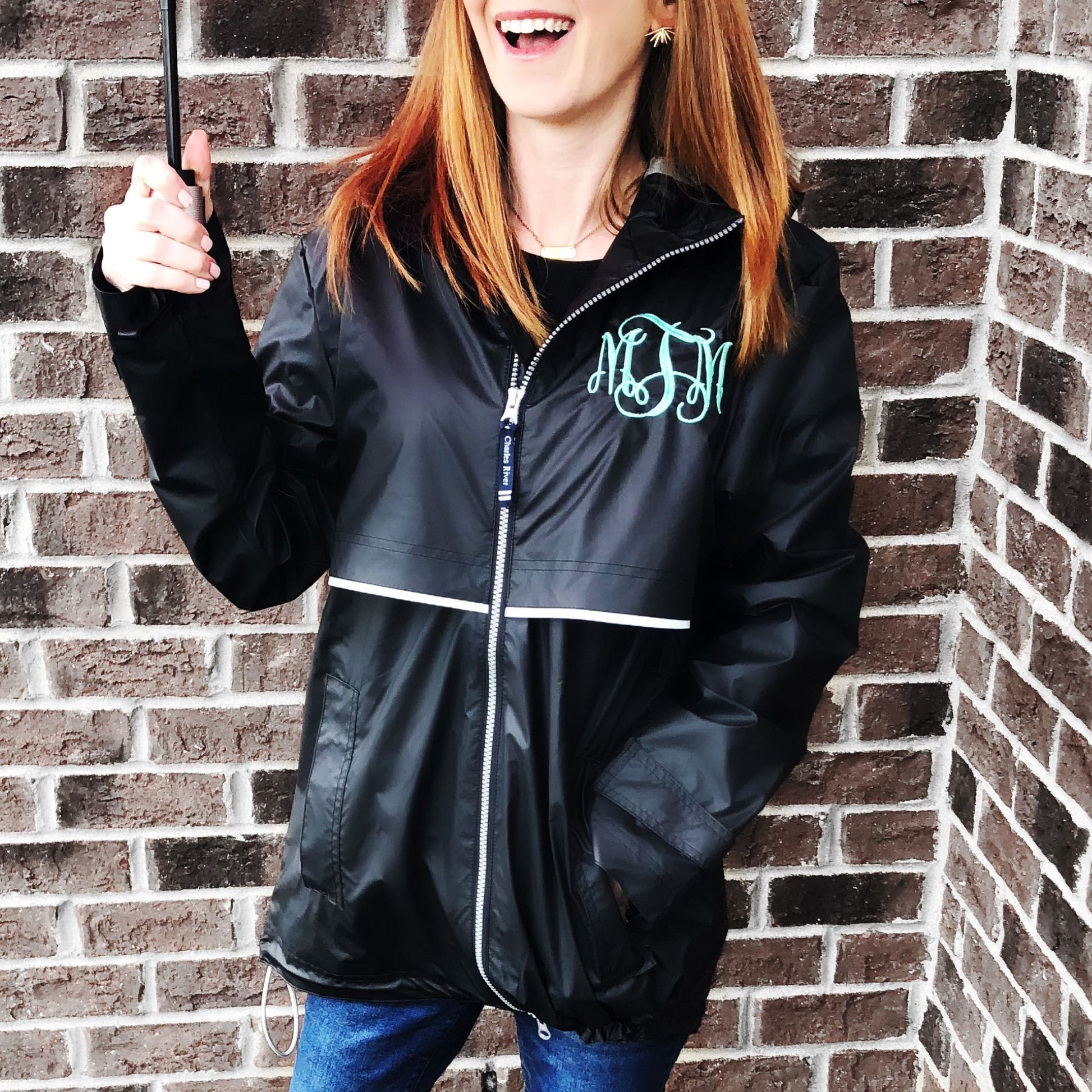 woman standing outside holding an umbrella wearing a black hooded full zip rain coat with custom monogram embroidered on the left chest in aqua thread.