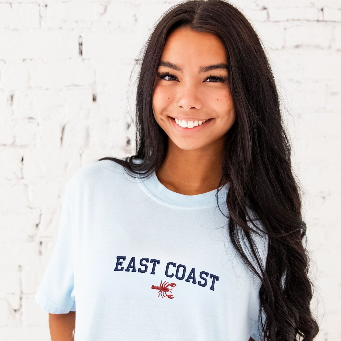Young woman wearing a light blue 'East Coast' t-shirt with a red lobster graphic against a white brick wall.