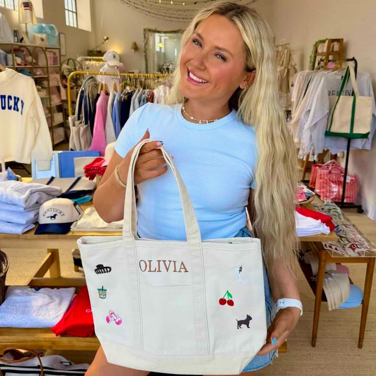 Woman holding a tote bag with 'OLIVIA' embroidered on it in a store setting.