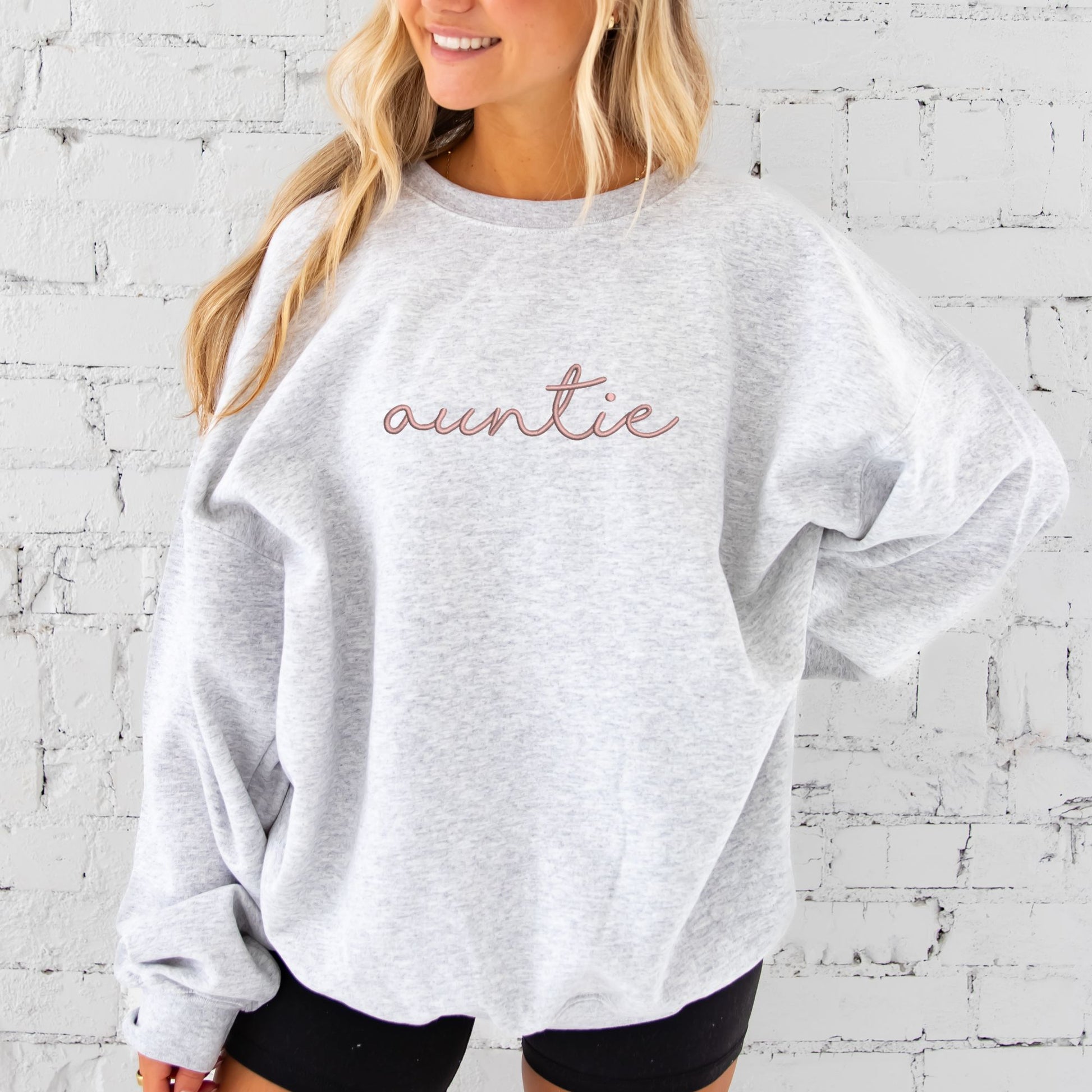 young woman wearing an oversized ash crewneck sweatshirt with embroidered auntie in lowercase script font across the chest in mauve thread