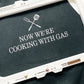 Charcoal apron with 'Now we're cooking with gas' embroidered text and crossed grilling utensils design