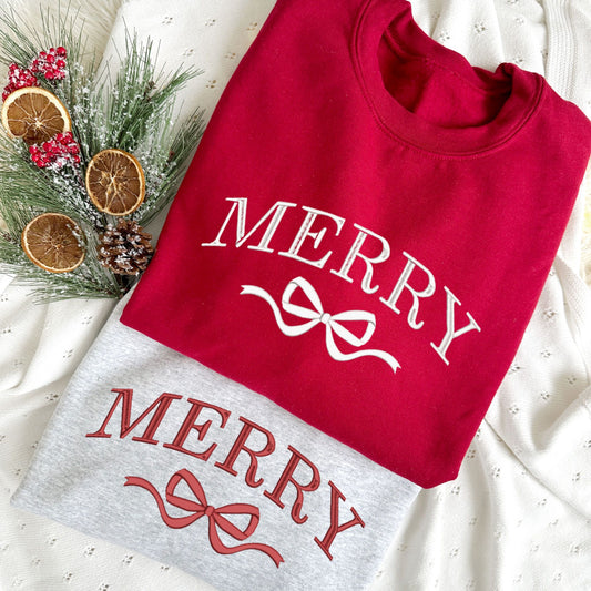 Christmas styled flat lay of a cardinal red crewneck with embroidered merry and bow design in white  thread and an ash crewneck with merry and bow design in red thread