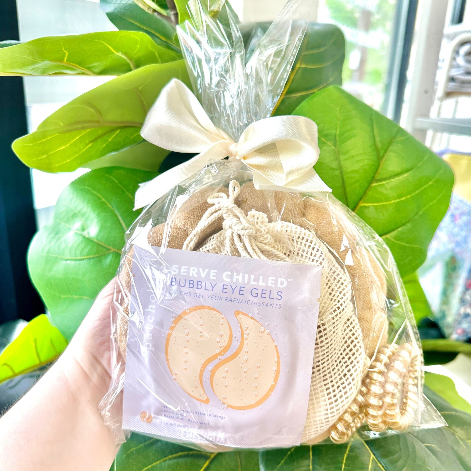 Packaged eye gels, reusable cotton rounds, spa head band, and coiled hair ties in shades of beige in a clear bag and tied with a decorative bow held in front of green leaves