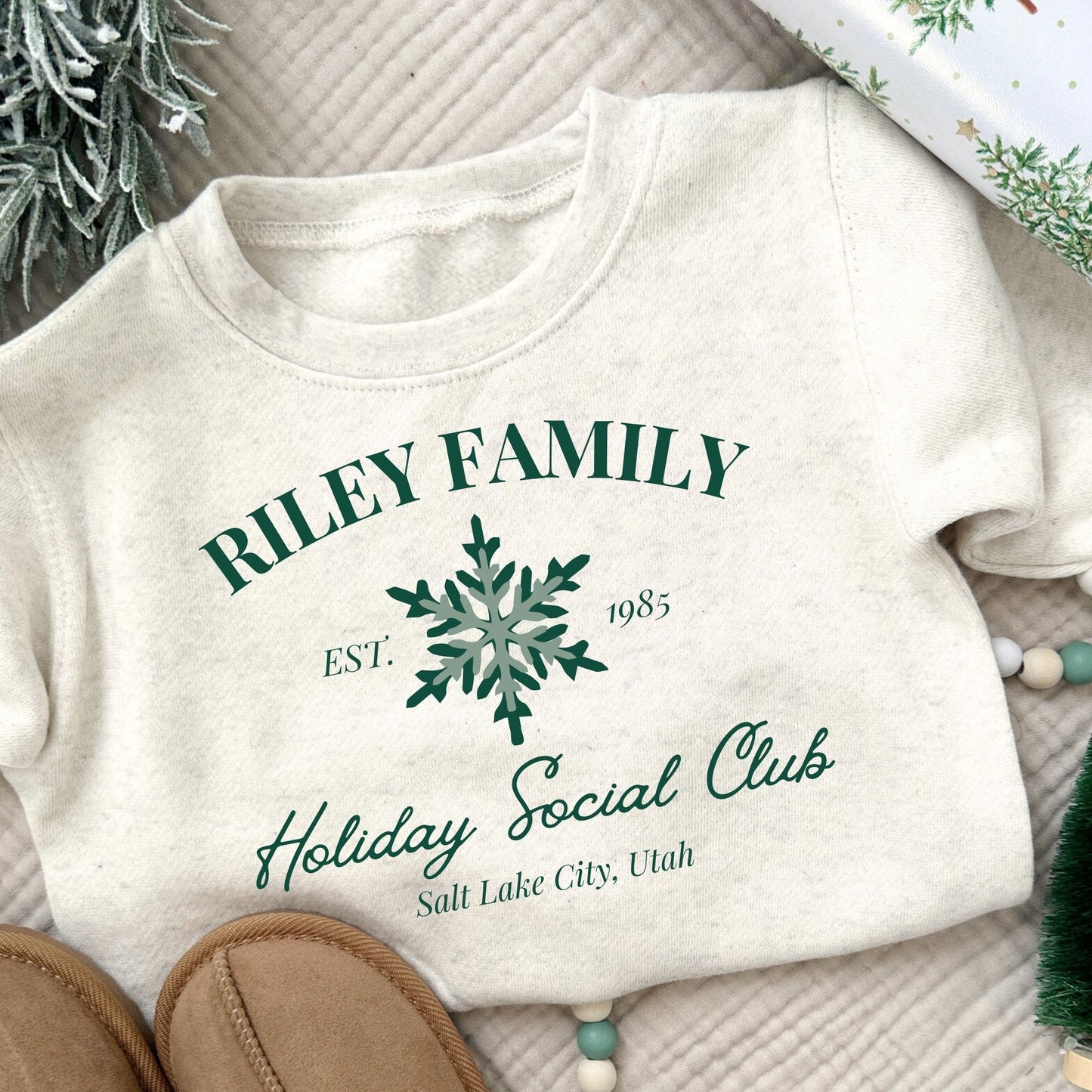 Natural Heather toddler crew with a custom print featuring a family name, Est date, snowflake, holiday social club, and location in green