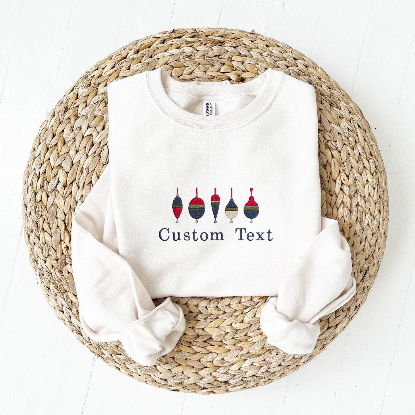 A white crewneck sweatshirt laid flat on a round woven straw mat, with rolled sleeves and embroidered design of six colorful fishing buoys above the words 'Custom Text'.