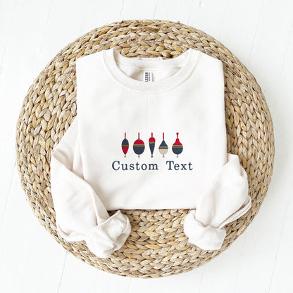 A white crewneck sweatshirt laid flat on a round woven straw mat, with rolled sleeves and embroidered design of six colorful fishing buoys above the words 'Custom Text'.