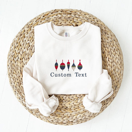 A white crewneck sweatshirt laid flat on a round woven straw mat, with rolled sleeves and embroidered design of six colorful fishing buoys above the words 'Custom Text'.
