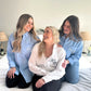 Three women wearing matching light blue shirts with monogrammed names on a bed.