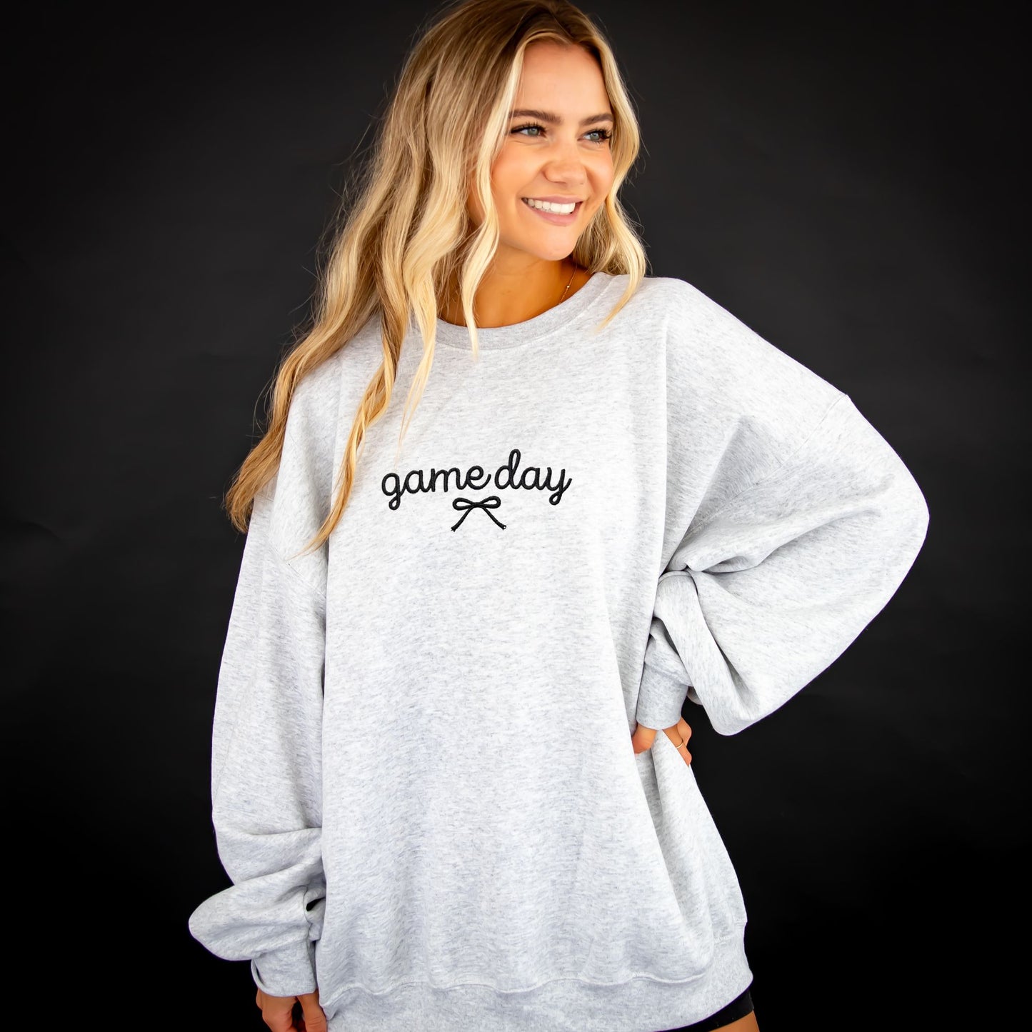 A woman with blonde hair wearing a light gray oversized crewneck sweatshirt with 'gameday' embroidered in black script across the chest, posed against a dark studio background.