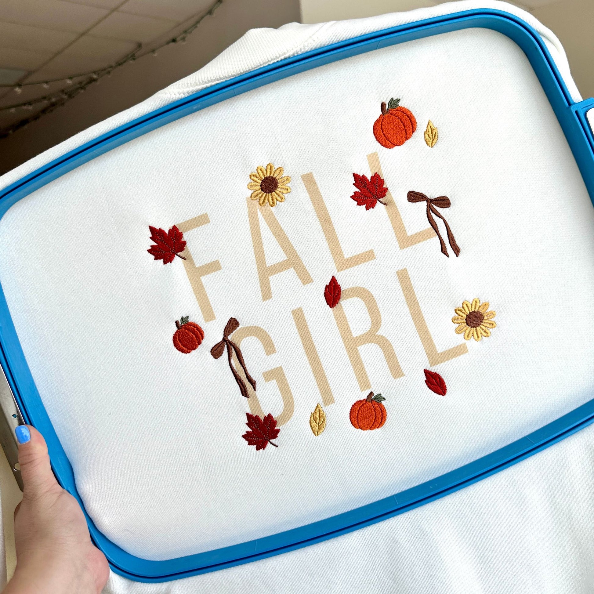 A close-up of a white fabric item inside a blue embroidery hoop, featuring the phrase 'FALL GIRL' in tan letters with autumn-themed embroidery including pumpkins, sunflowers, leaves, and bows. A hand with blue nail polish is holding the hoop.