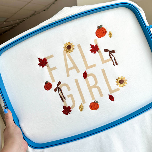 A close-up of a white fabric item inside a blue embroidery hoop, featuring the phrase 'FALL GIRL' in tan letters with autumn-themed embroidery including pumpkins, sunflowers, leaves, and bows. A hand with blue nail polish is holding the hoop.