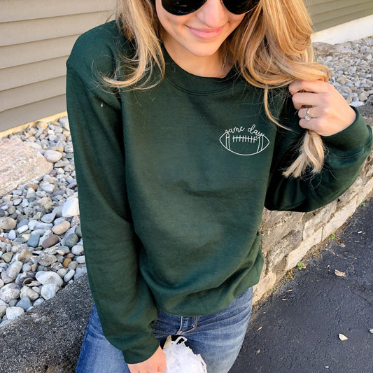 woman wearing a forest green crewneck sweatshirt with an embroidered outline game day design in white thread on the left chest