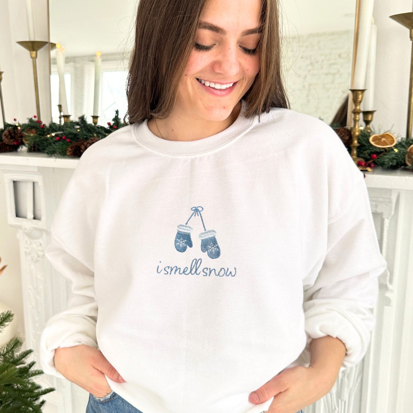 young woman wearing a white crewneck sweatshirt with i smell snow and mittens embroidered on the center chest in powder blue thread