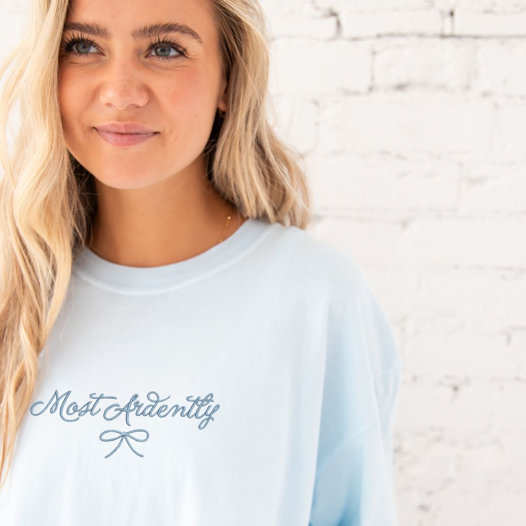 close up of a young woman wearing a chambray comfort colors t-shirt with embroidered most ardently and bow design in baby blue thread