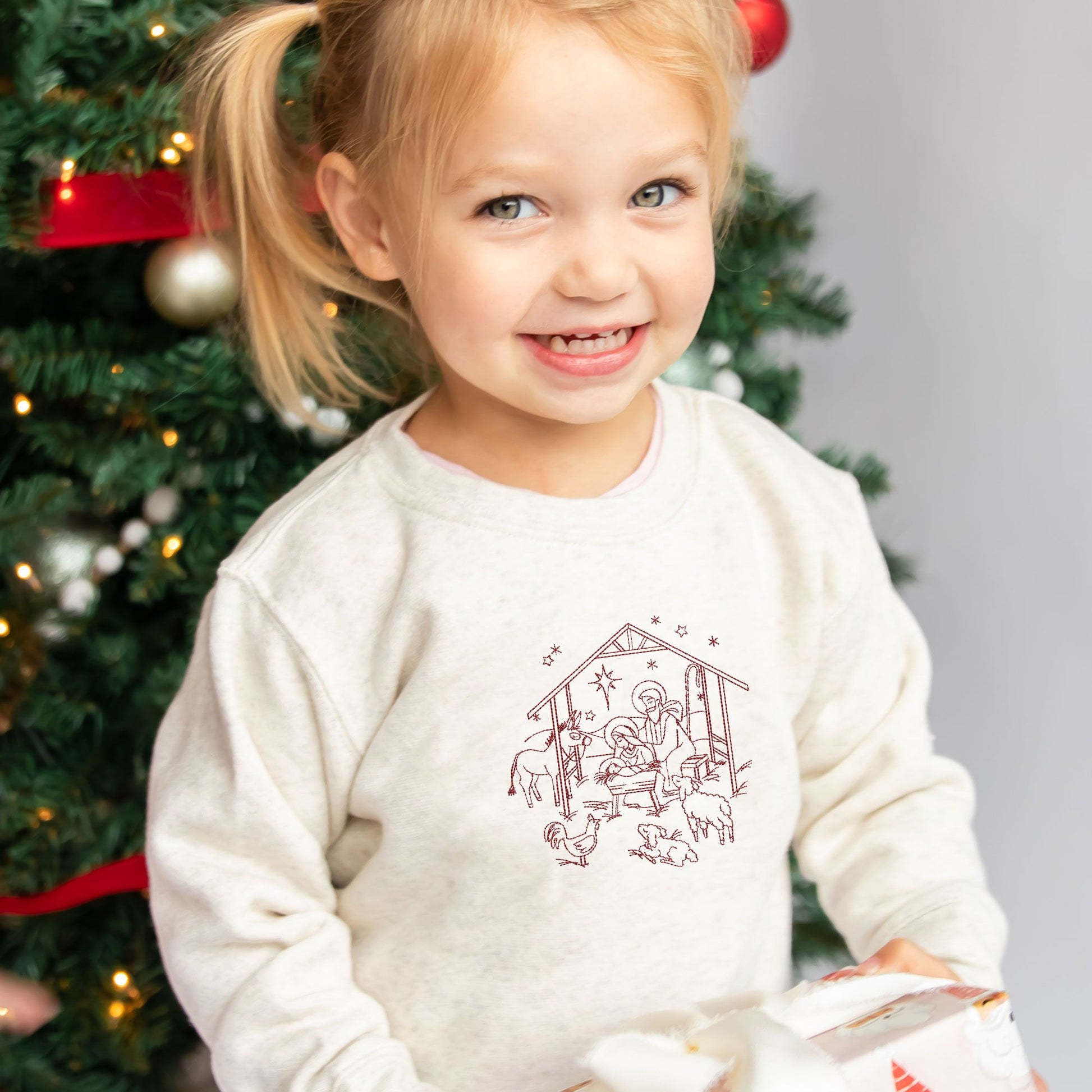 toddler girl wearing  heather oatmeal crewneck with embroidered stitched nativity scene in red thread