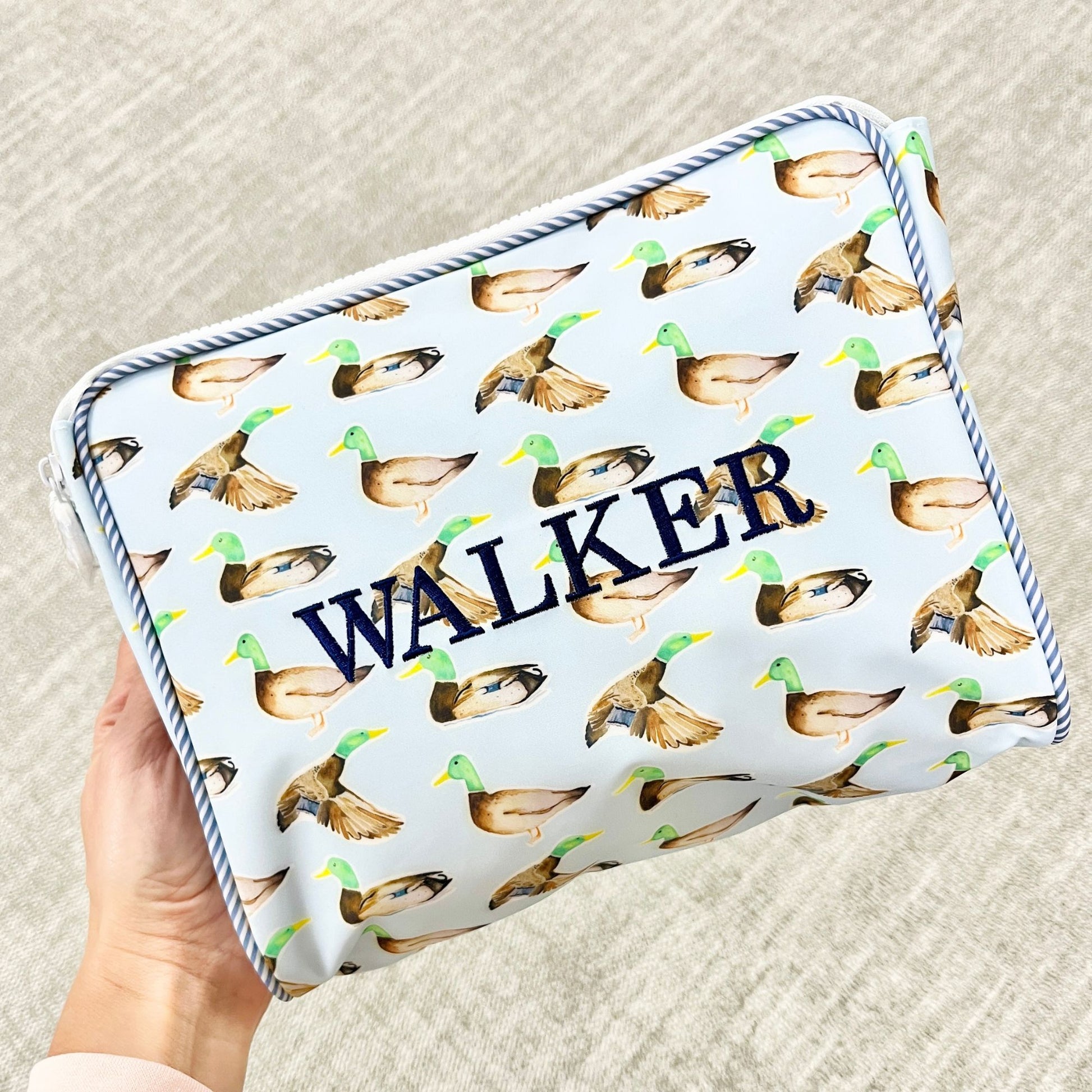 Pouch with duck pattern and 'WALKER' text embroidered held by a hand on a textured surface