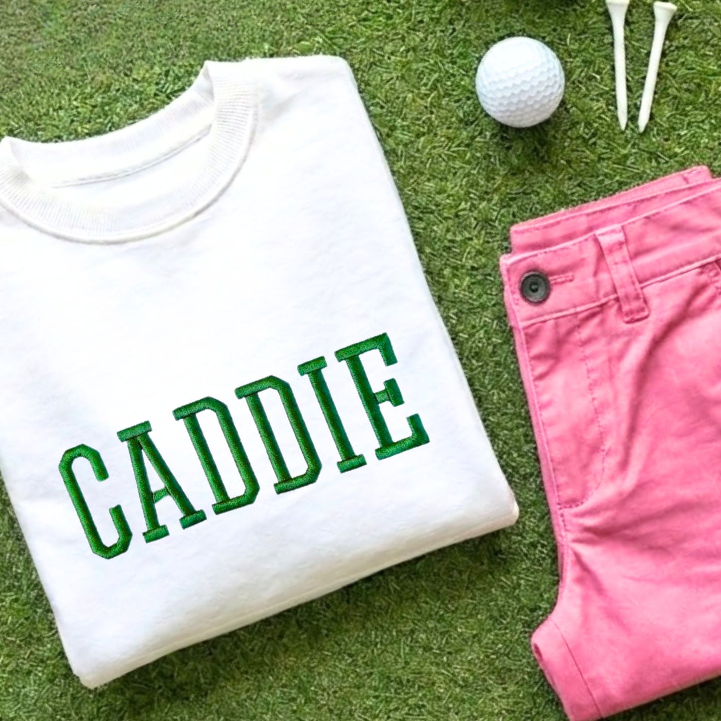 White sweatshirt with 3d embroidered 'CADDIE' design in kelly green thread, paired with pink shorts, a golf ball and golf tees