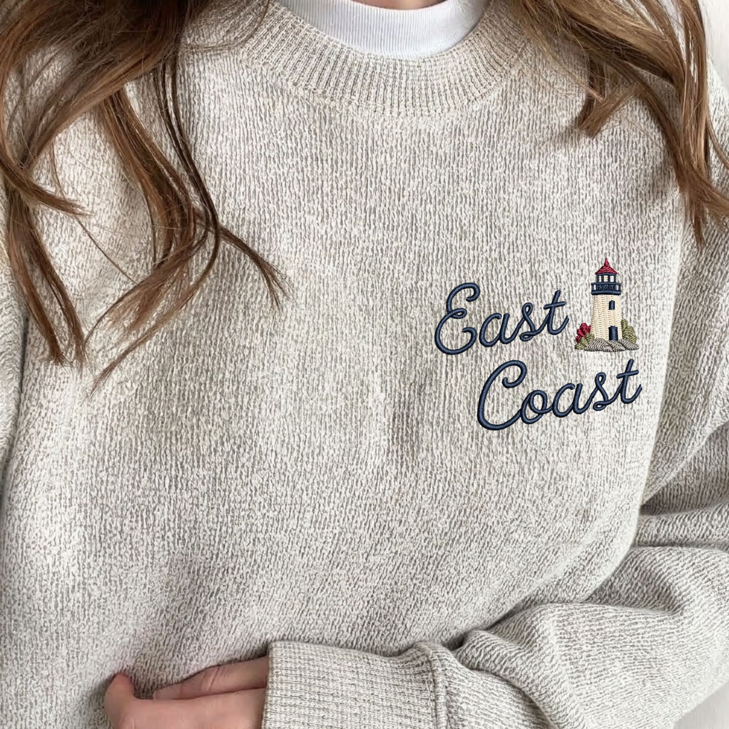 A close-up of a person wearing an oatmeal heather knit sweater with navy blue 'East Coast' script embroidery and a small lighthouse graphic on the chest.