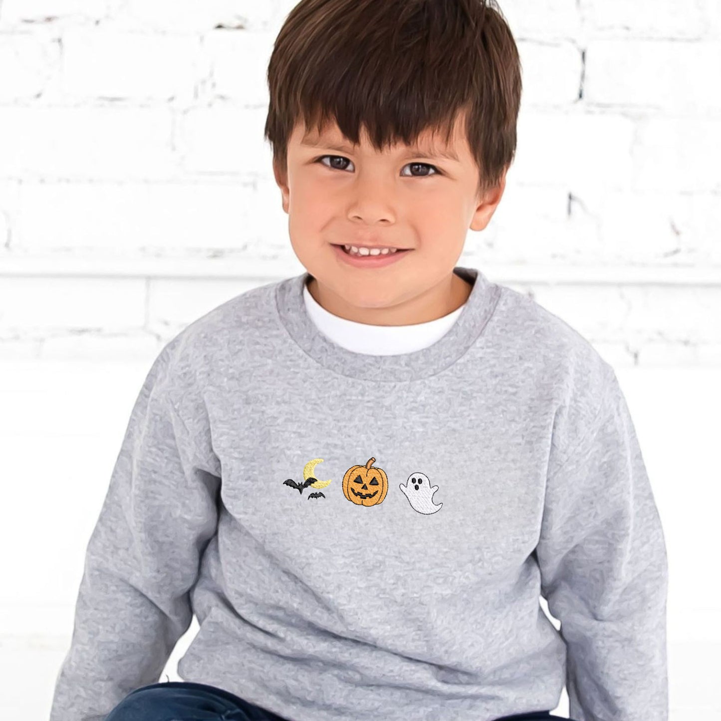 Little boy wearing a gray sweatshirt with Halloween-themed embroidered icons (moon/bats, jack-o-lantern, and ghost) on the center chest. He's sitting on the ground and there's a white brick wall behind him.