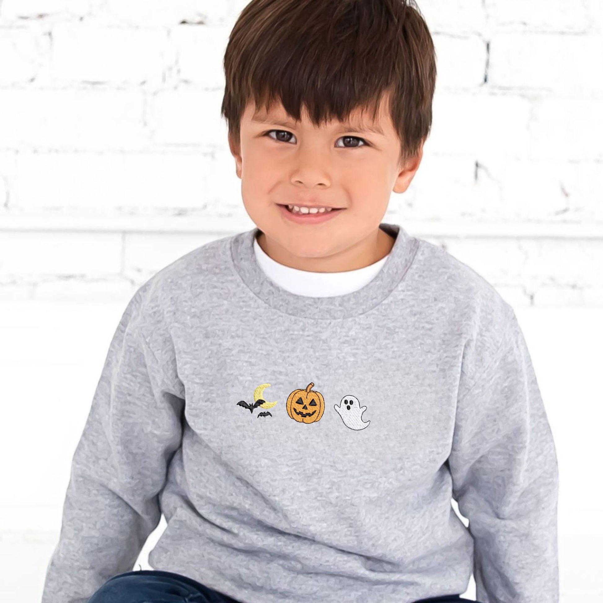 Little boy wearing a gray sweatshirt with Halloween-themed embroidered icons (moon/bats, jack-o-lantern, and ghost) on the center chest. He's sitting on the ground and there's a white brick wall behind him.