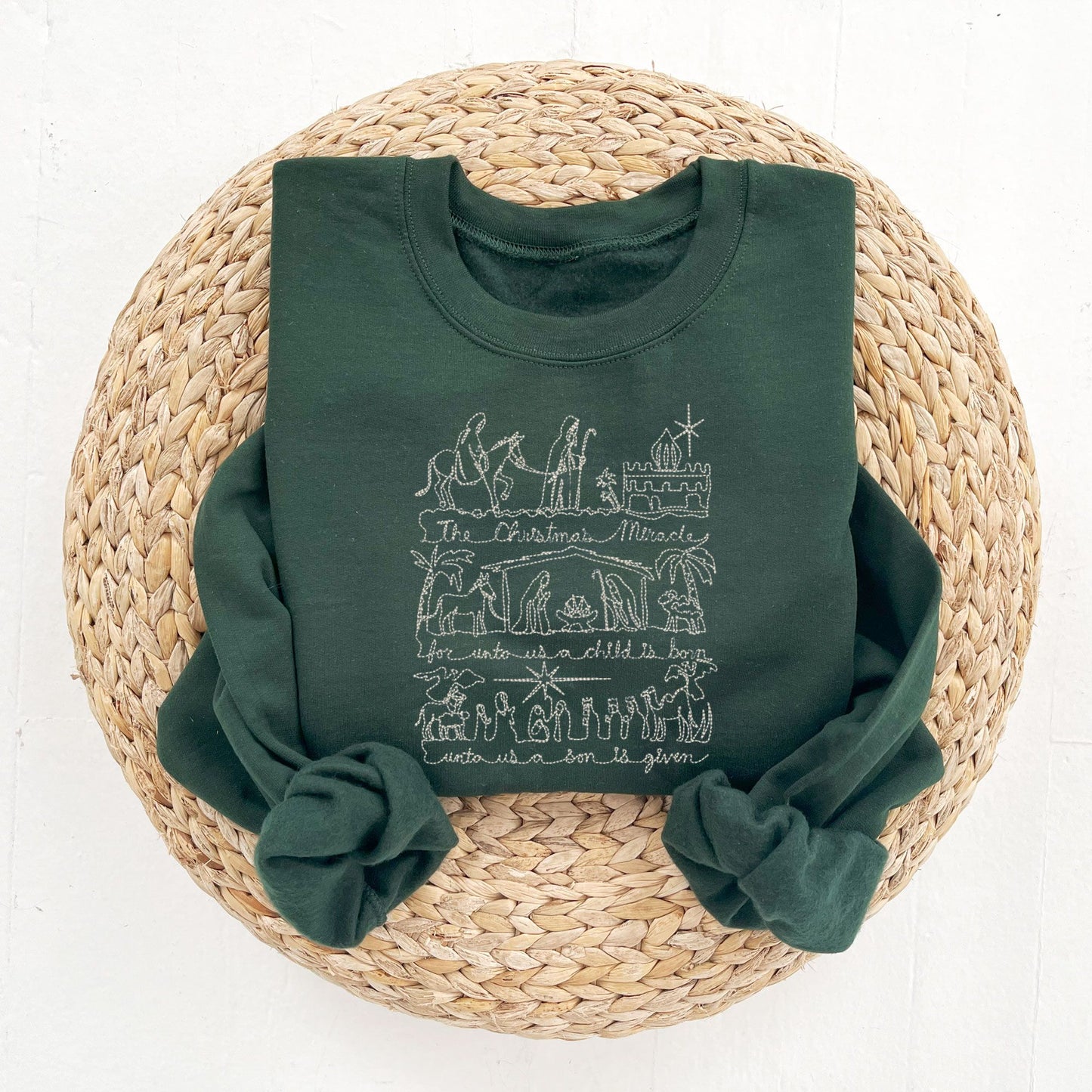 flat lay photo of a forest green crewneck sweatshirt with embroidered stitched drawing of the Christmas nativity in natural thread.