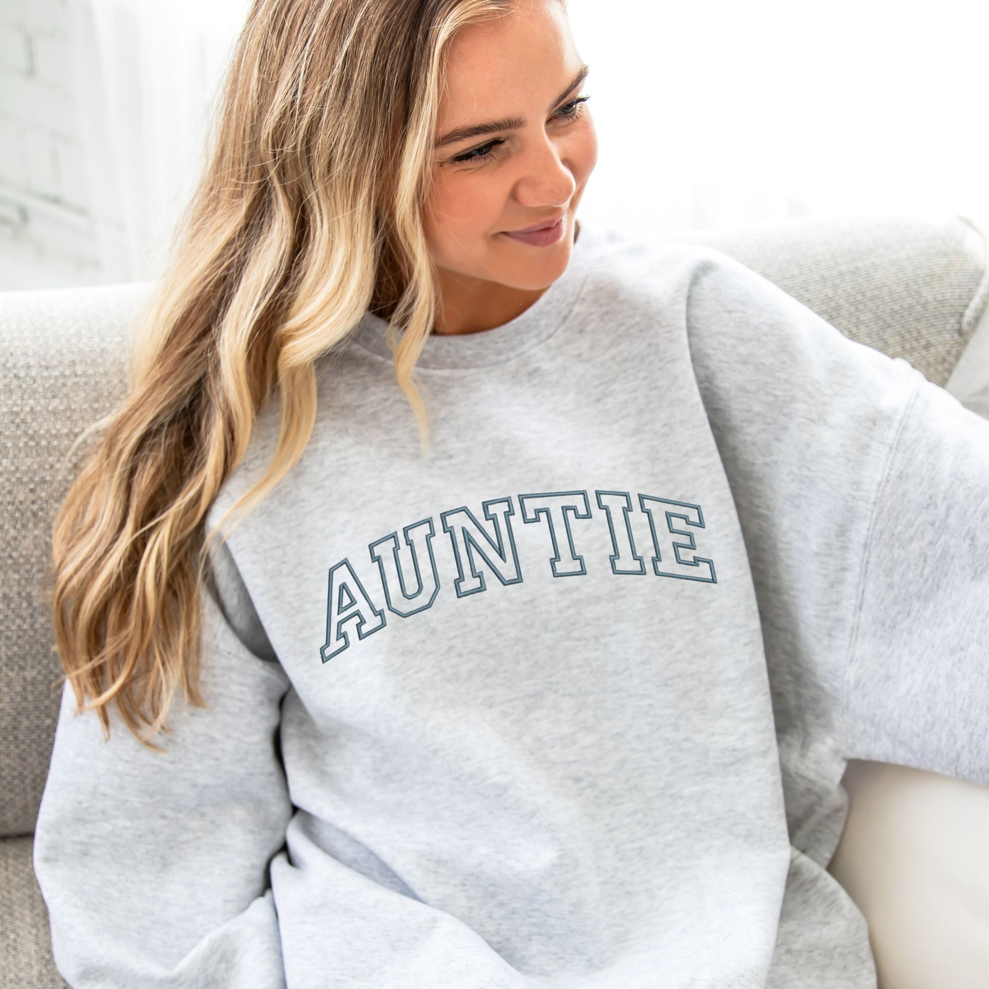 young woman wearing an oversized ash crewneck sweatshirt with embroidered AUNTIE in all caps athletic block font across the chest in french blue thread