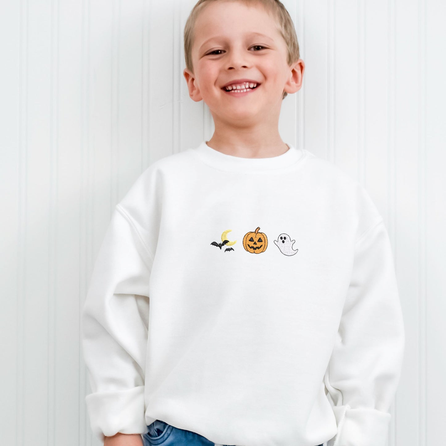 Little boy wearing a white sweatshirt with Halloween-themed embroidered icons (bats/moon, jack o lantern, ghost). He's standing in front of a white wall