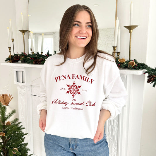 young woman wearing a white crewneck sweatshirt with a custom print featuring a family name, Est date, snowflake, holiday social club, and location in red