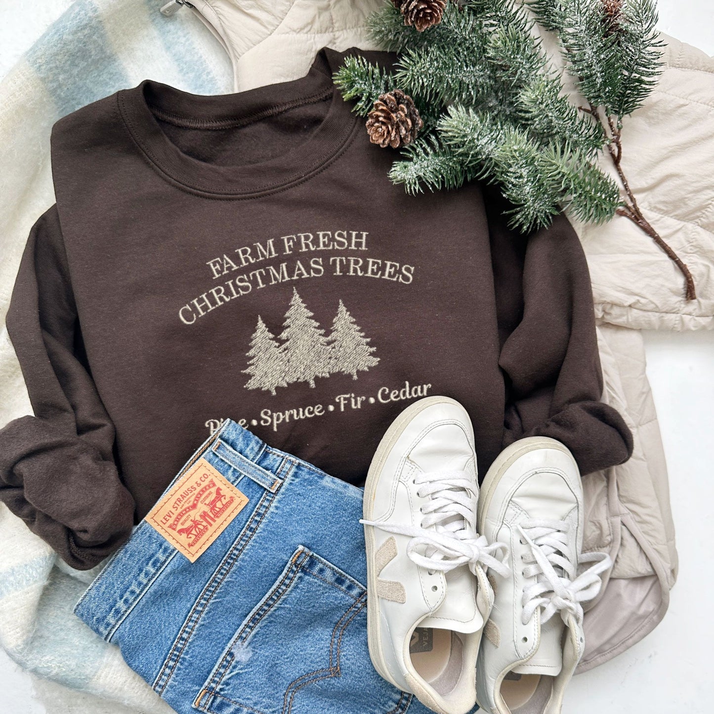 Christmas styled flat lay of a dark chocolate crewneck sweatshirt with embroidered farm fresh Christmas trees design with Christmas tree embroidery across the chest in natural green thread