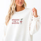 woman wearing a sweet cream crewneck sweatshirt with embroidered merrier with a martini text in red thread and martini glass design with bow and pine sprig across the chest