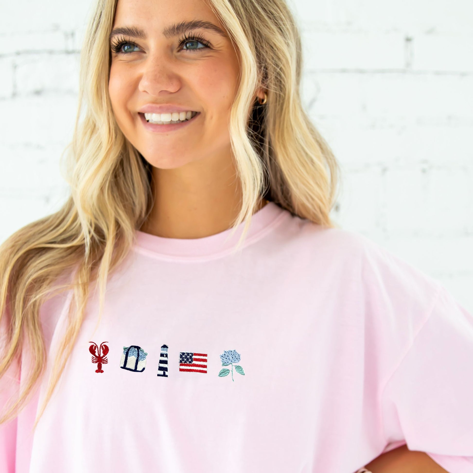 Woman wearing a pink t-shirt with embroidered designs on a white brick wall background
